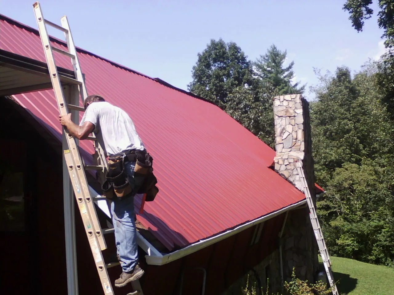 Roofer on a ladder beside a red metal roof and stone chimney — Soffit & Fascia Repair by Roofing Brothers in West Salisbury